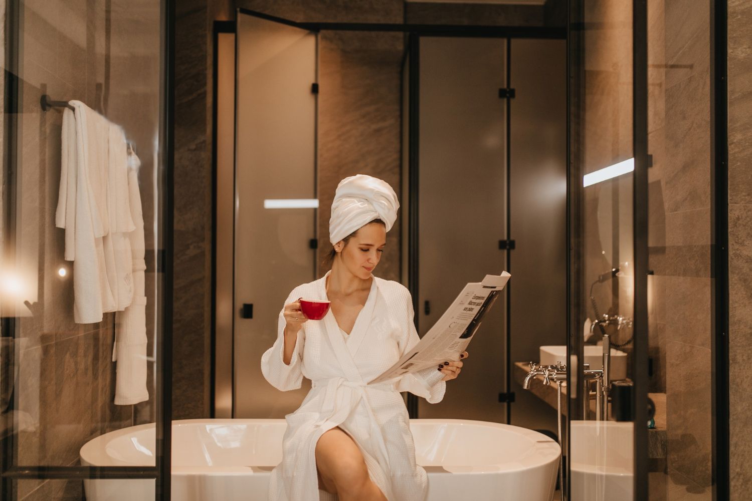 A woman in a bathrobe is sitting on a bathtub reading a newspaper.