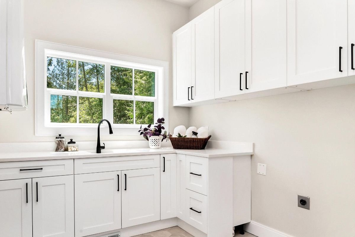 A laundry room with white cabinets , a sink , and a window.