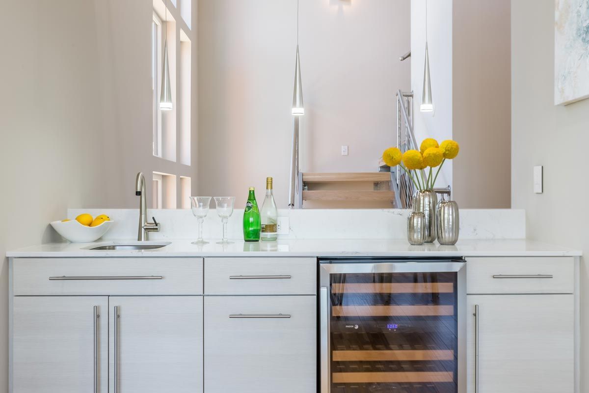 A kitchen with white cabinets , a wine cooler , a sink and a mirror.