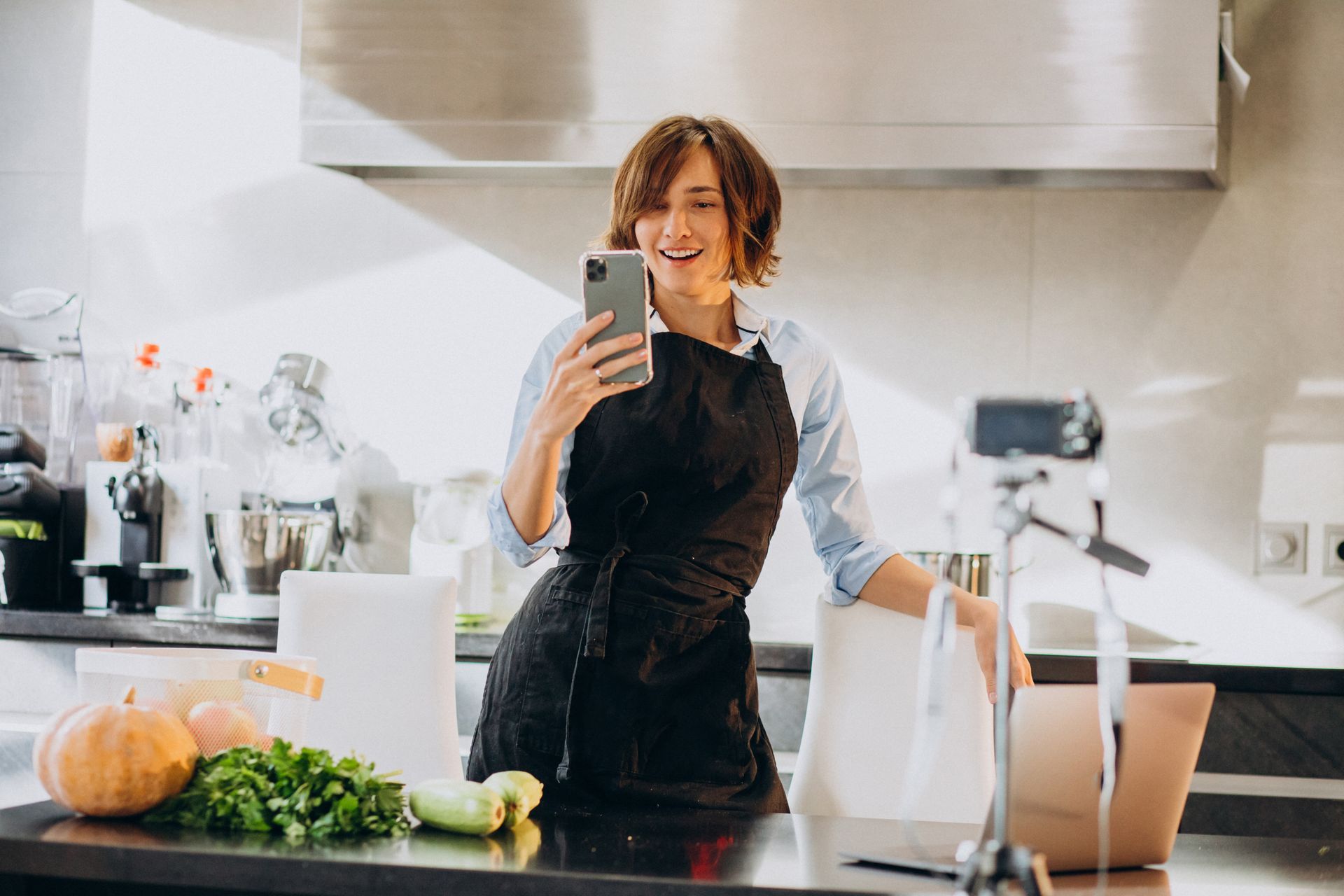 A woman in an apron is taking a selfie in a kitchen.