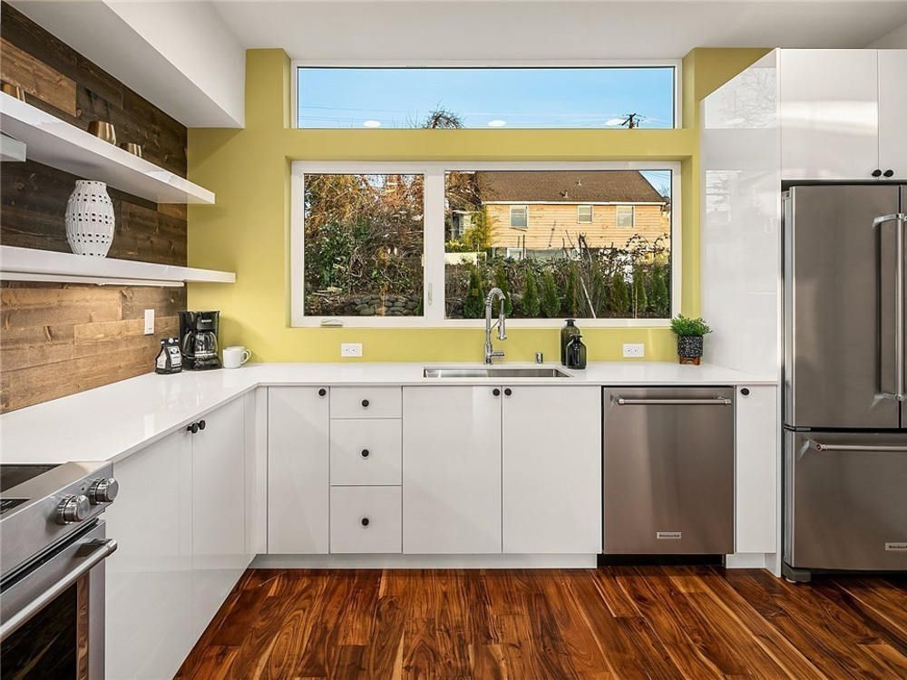 A kitchen with white cabinets , stainless steel appliances , hardwood floors and a large window.
