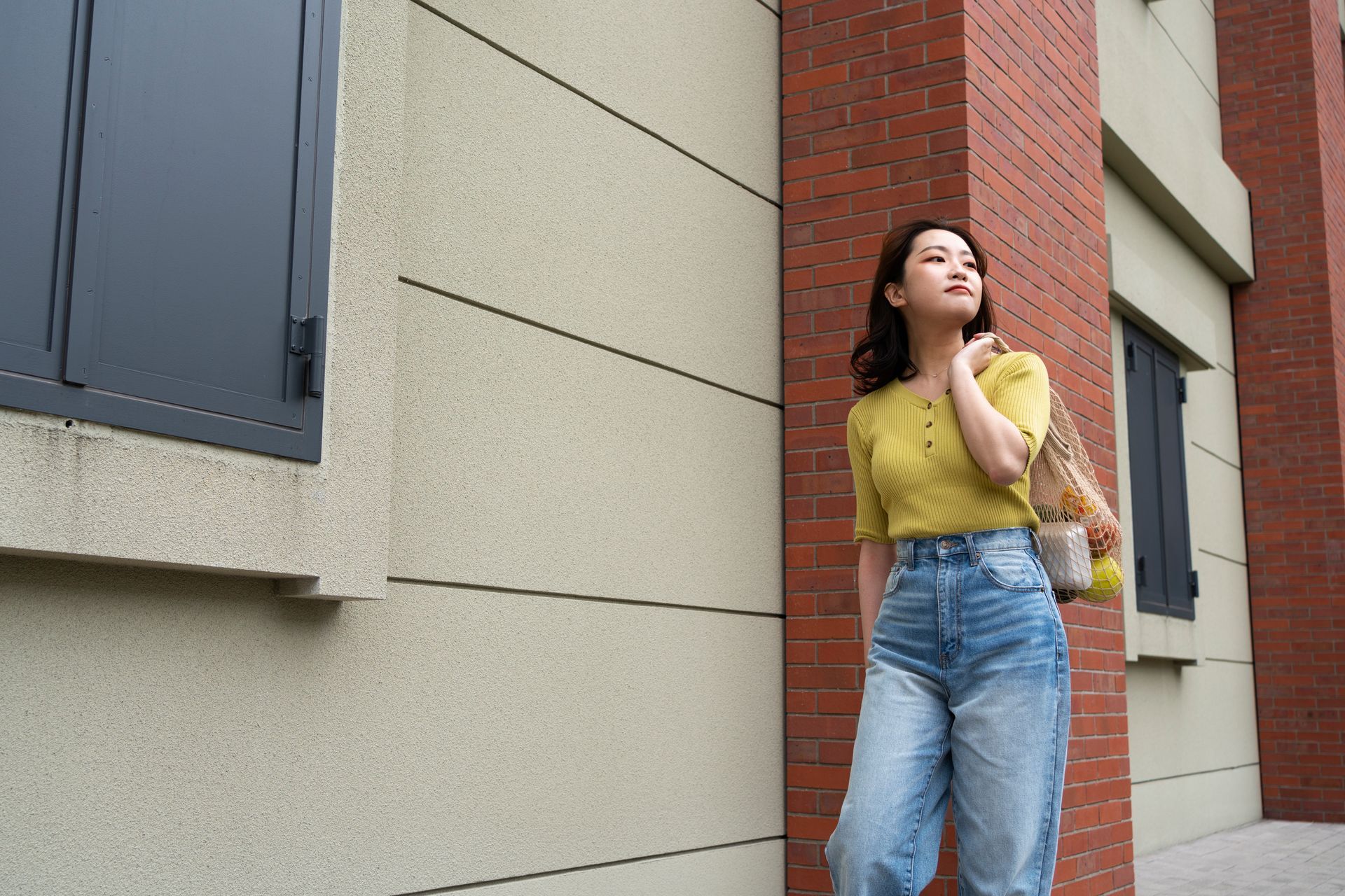 A woman in a yellow shirt and blue jeans is standing in front of a brick building.
