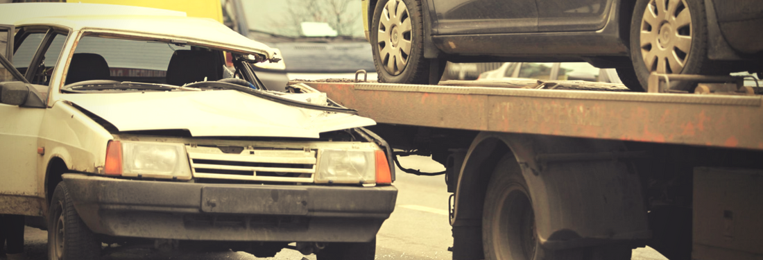 Damaged white sedan beside a tow truck on a road, with another car visible in the background