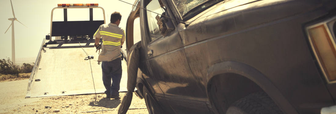 Worker in a yellow safety vest beside a dark car being towed in a dusty roadside scene