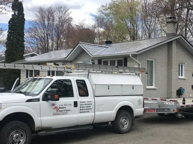 Un camion blanc est stationné devant une maison au toit en tôle. Une échelle se trouve sur le toit.