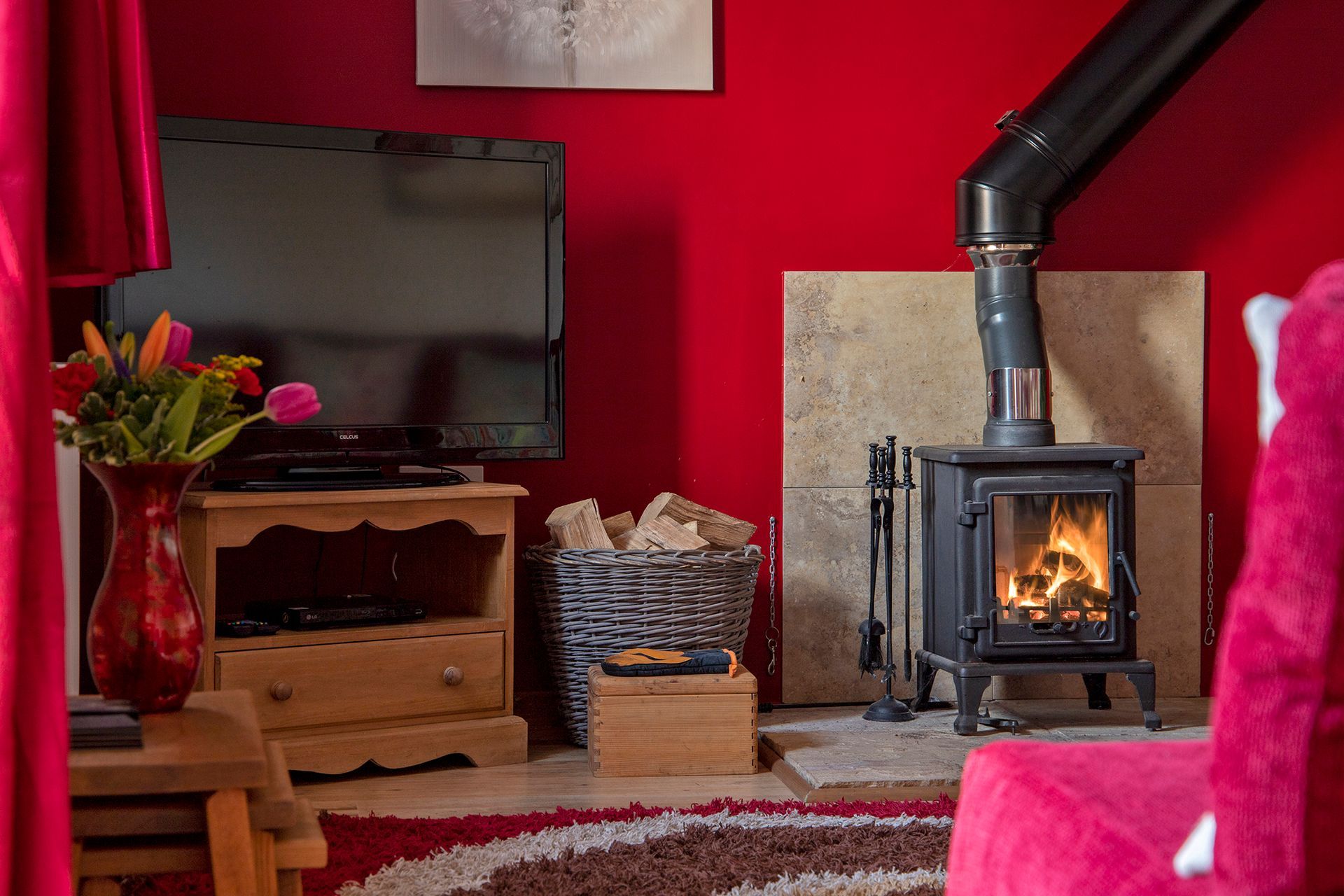 A lit wood burner with a basket of logs, TV and vase of flowers