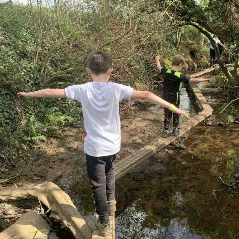 two boys balancing on a narrow wooden plank across a stream