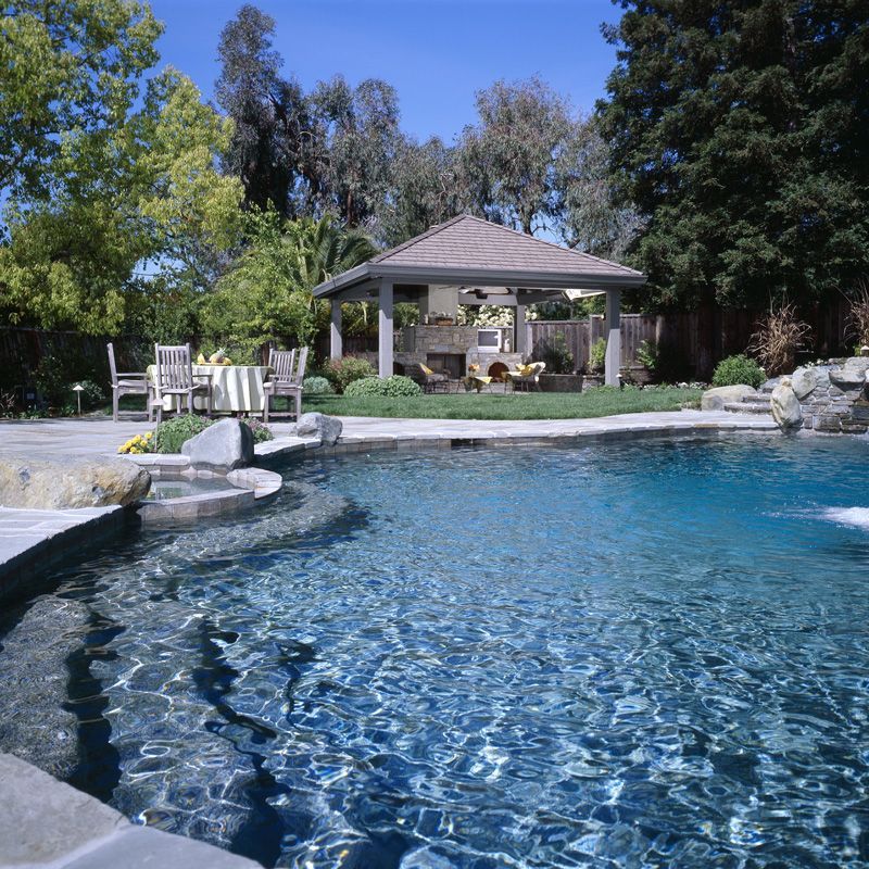 Backyard pool with clear blue water, stone edging, and a gazebo surrounded by trees.