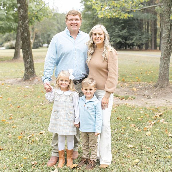 Family of four standing together outdoors in a park, smiling in soft autumn light.