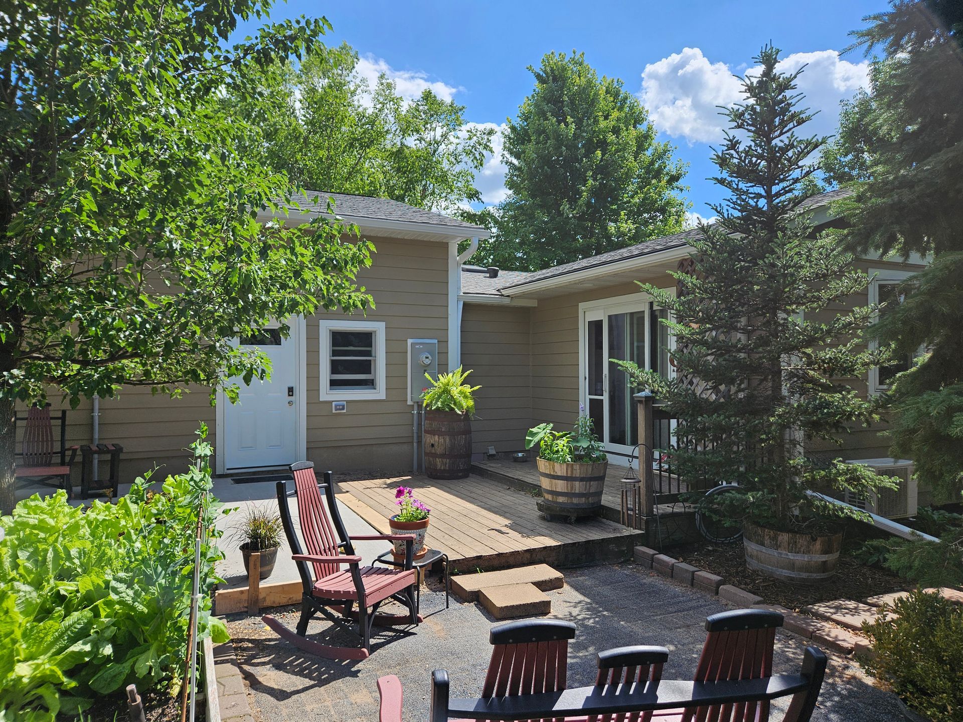 Backyard with a deck, chairs, trees, and a building with a white door and a window on a sunny day.