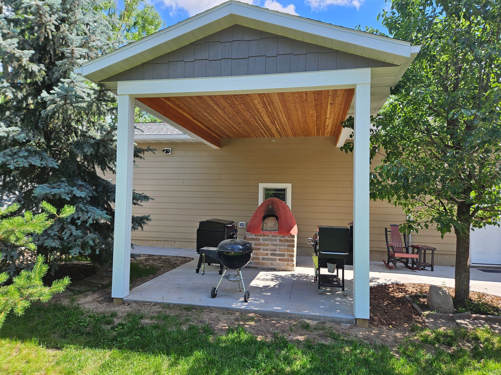 A patio shelter with grills and a brick structure, located on a concrete slab. Green grass and trees surround.
