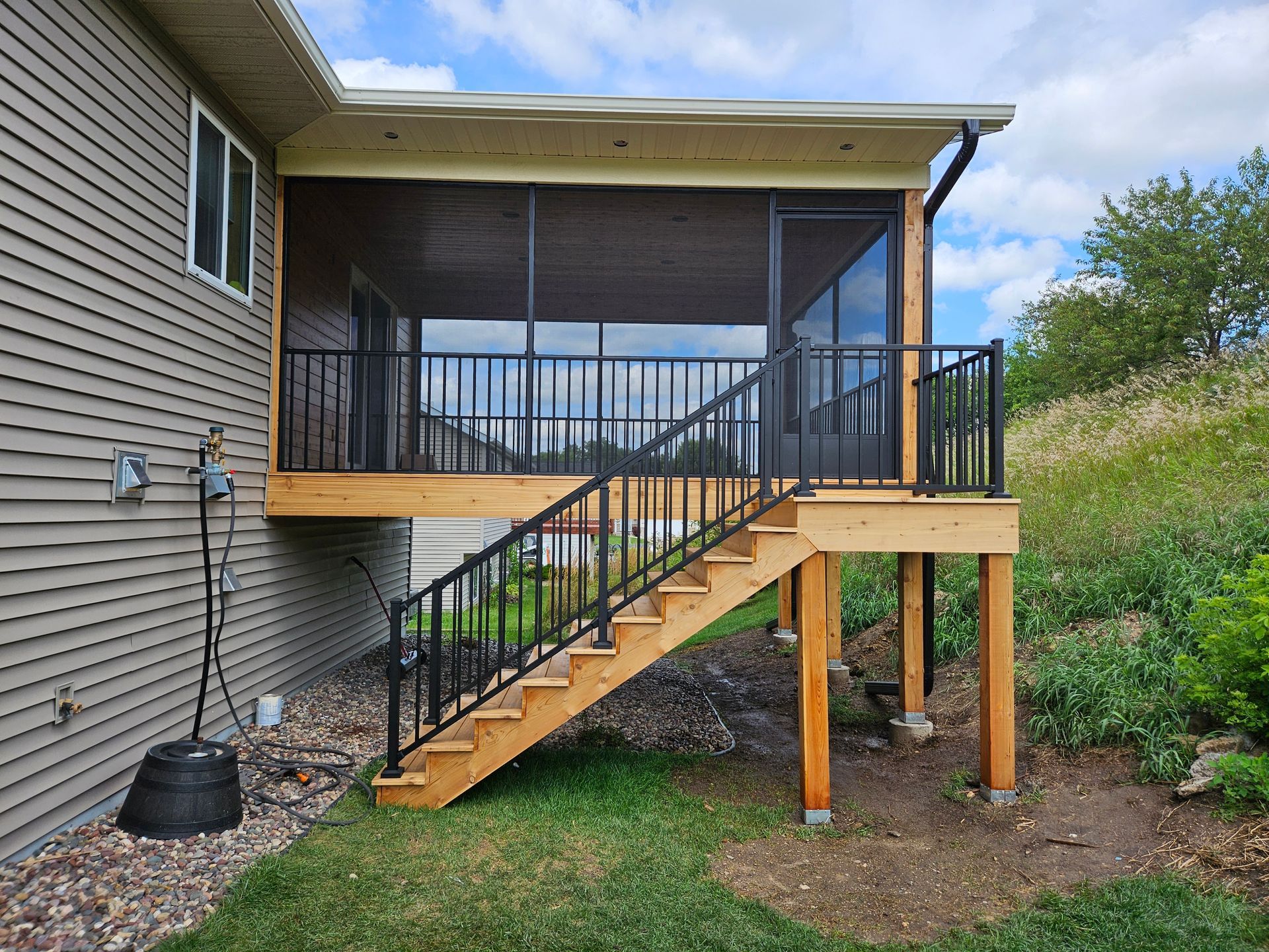 Screened-in porch with stairs, wooden deck, black railing, and siding, with a grassy hill in the background.