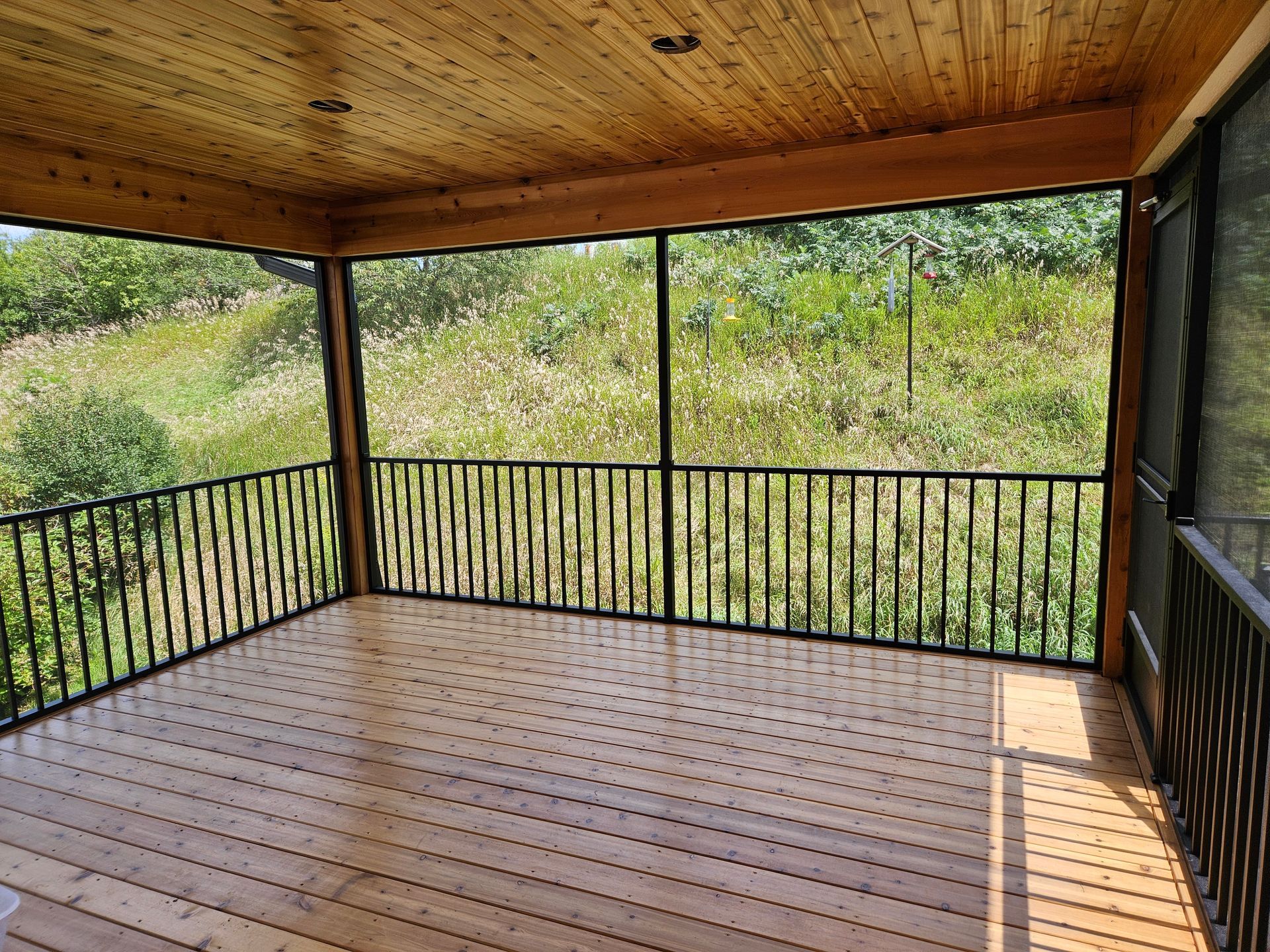 Screened porch with wood ceiling and floor, black railing, overlooking a grassy field.