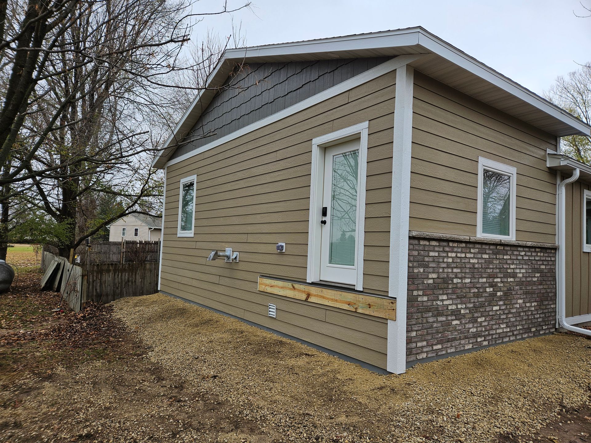 Tan and brown house with brick base, wooden door, and two windows.