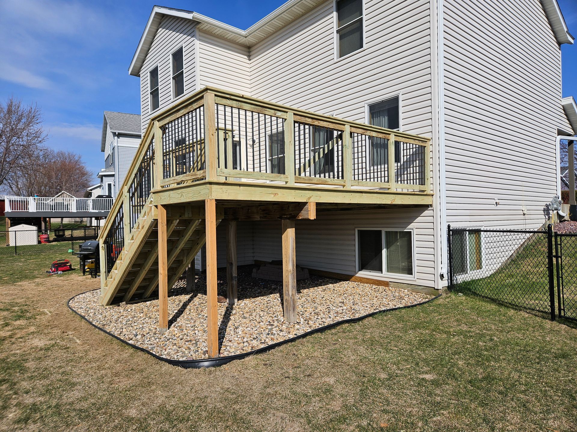 Wooden deck attached to a two-story house with stairs, supported by wooden beams, on a gravel bed.