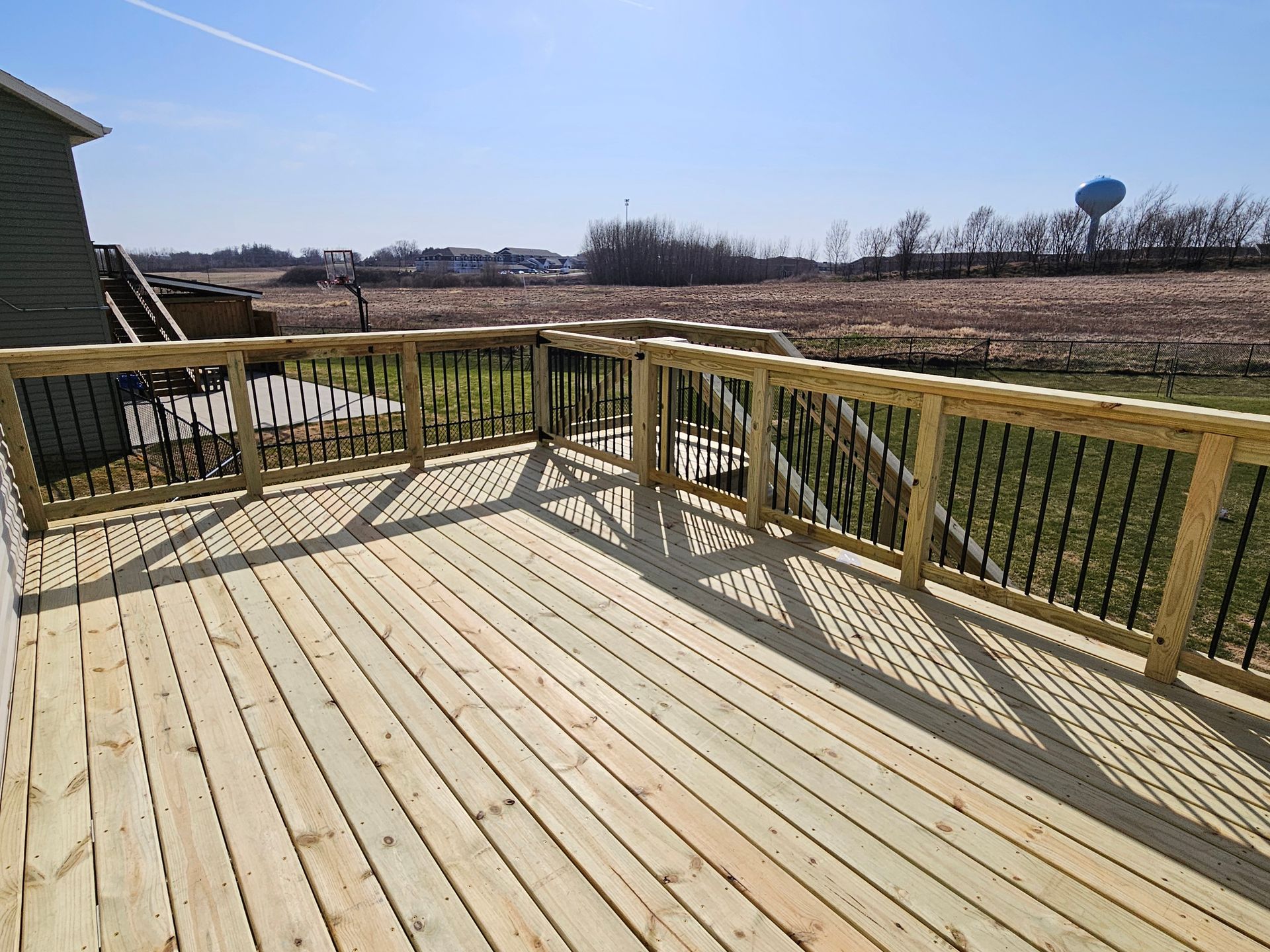 Wooden deck with black railing on a sunny day, overlooking a field and a water tower.