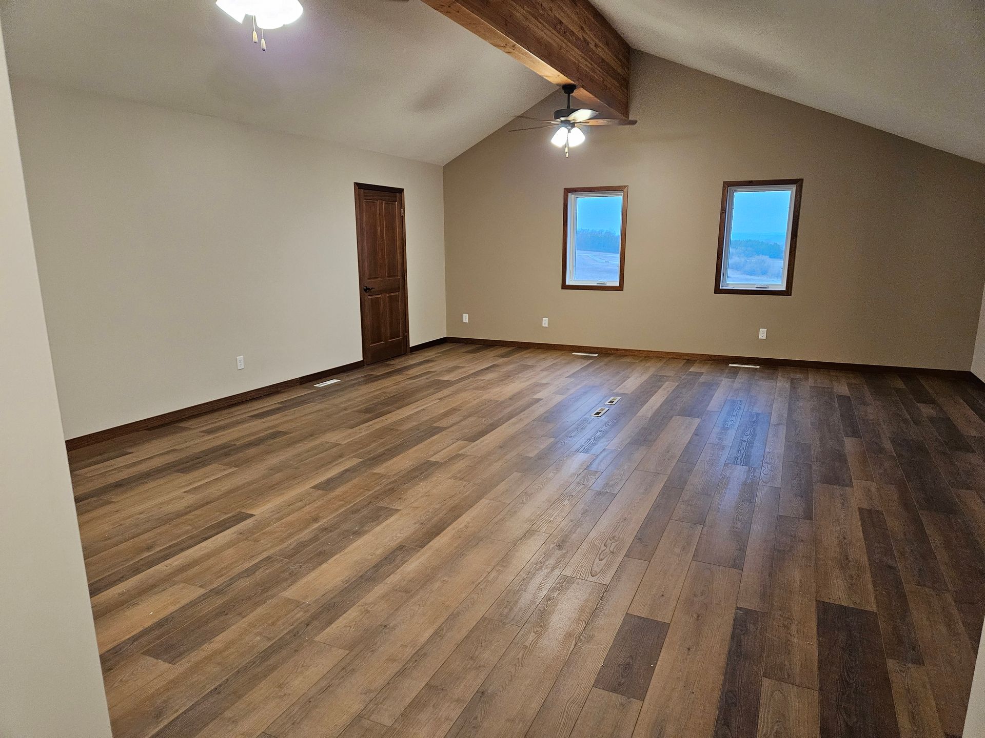 Empty room with brown hardwood floors, tan walls, two windows, and a wooden beam ceiling.