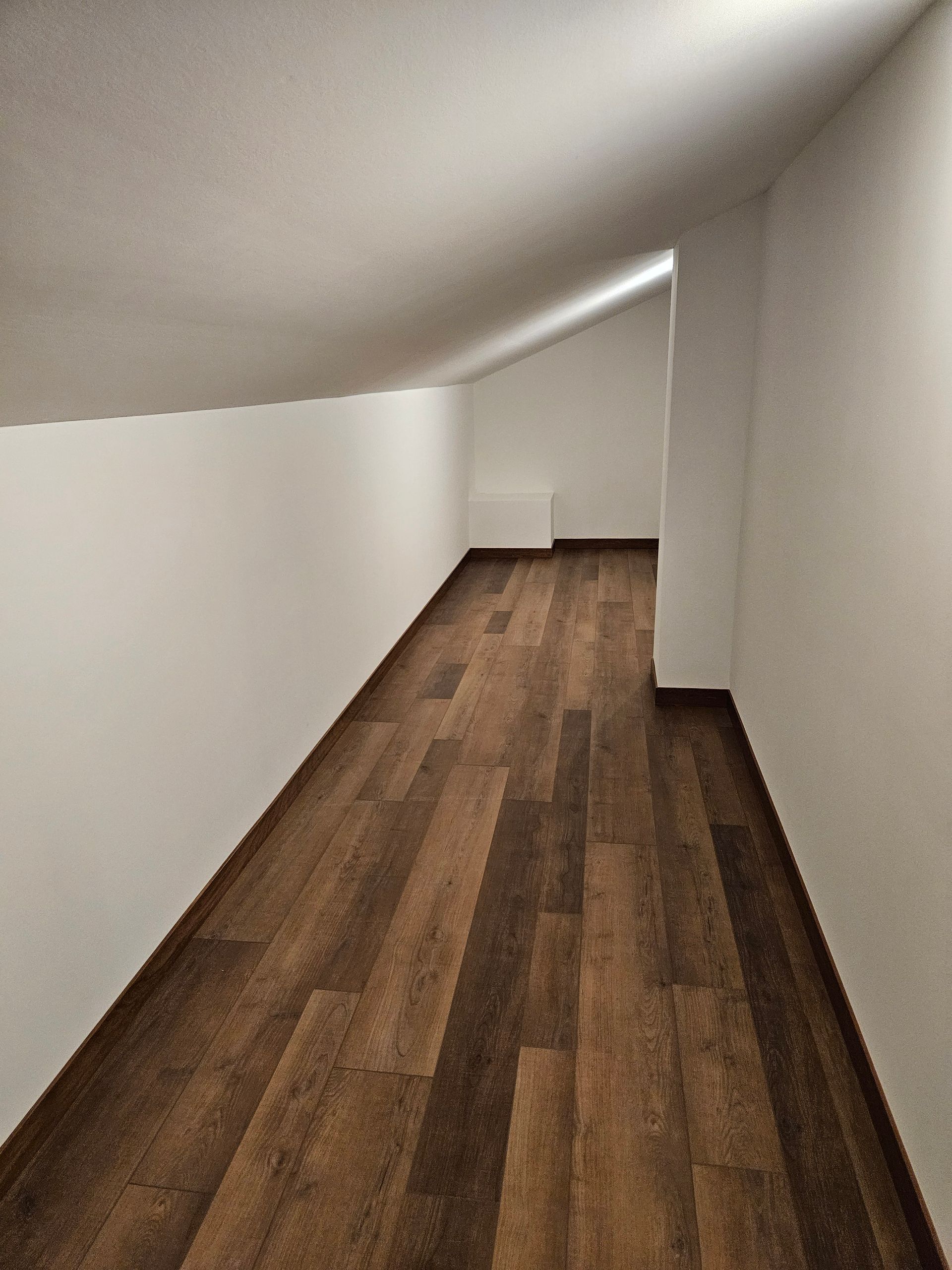 Narrow, sloping attic room with brown wood-look flooring and white walls.