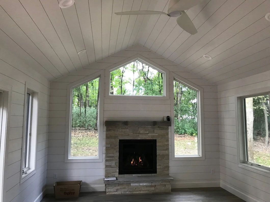 Interior of a white-paneled room with a stone fireplace and windows overlooking a green forest.