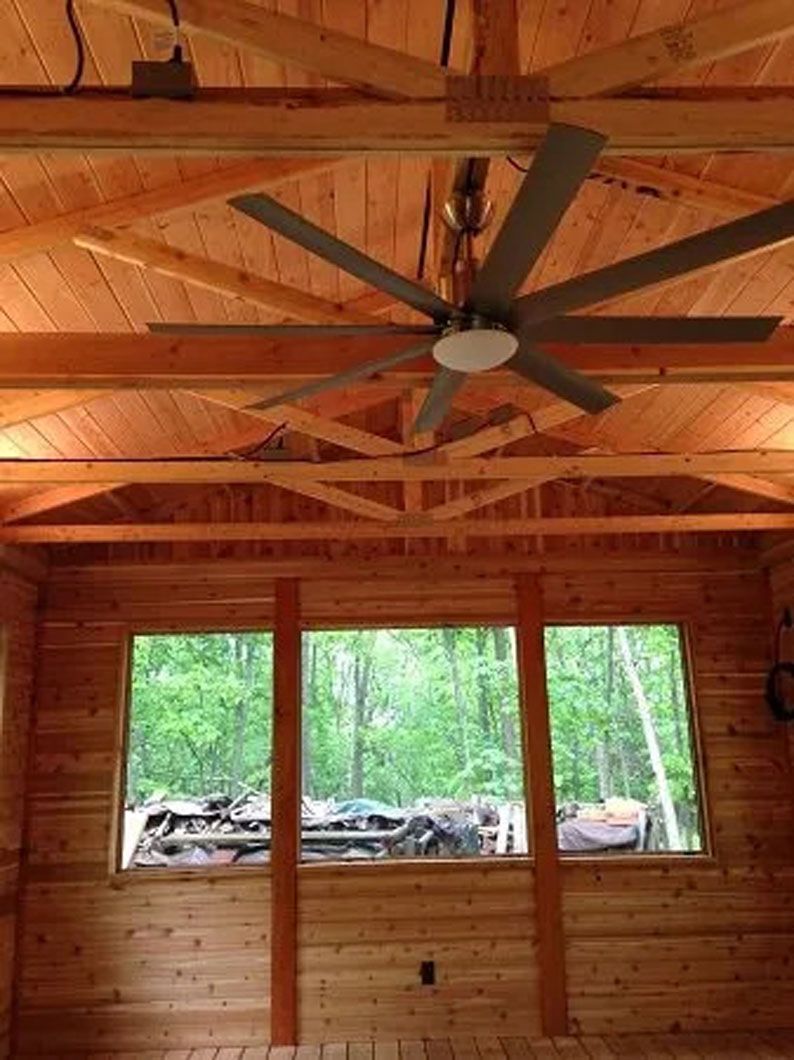 Wooden cabin interior with large ceiling fan and windows looking out to a wooded area.