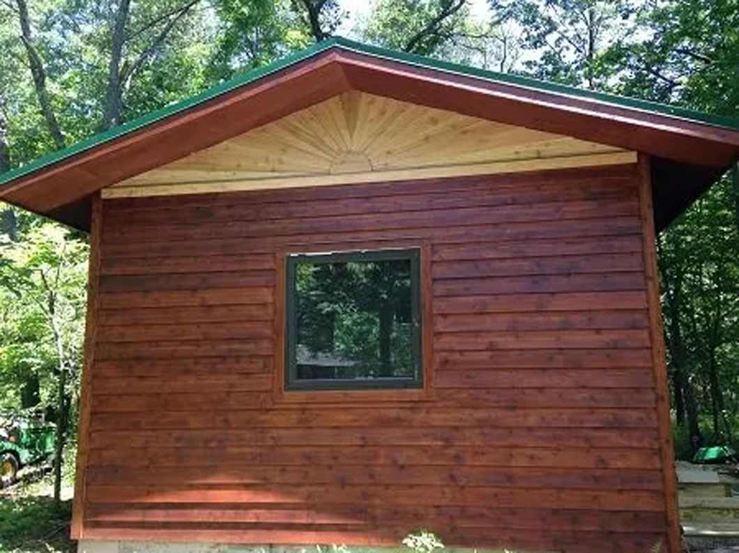Small brown cabin with a green roof and a square window surrounded by trees.