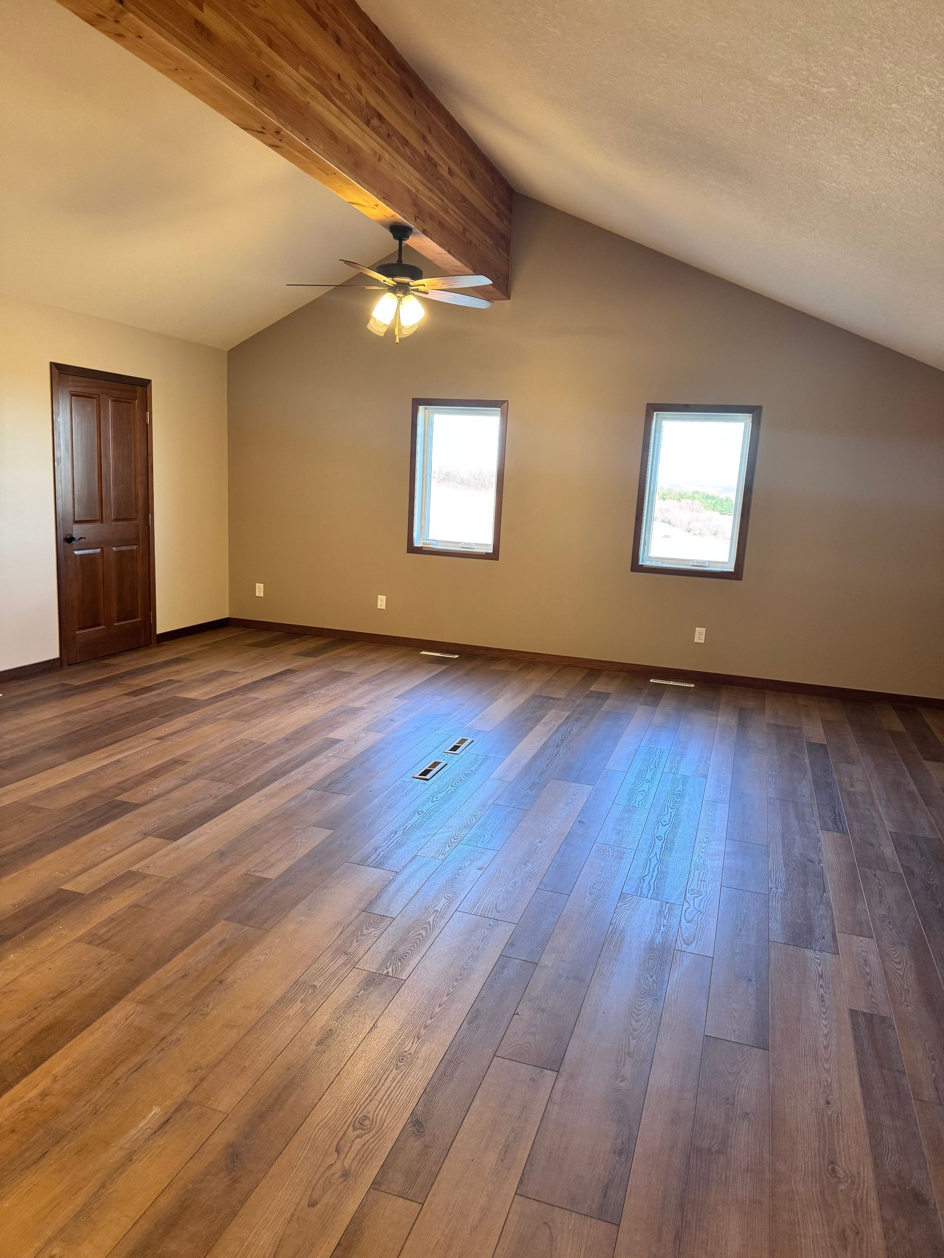 Spacious room with wood beam ceiling, wood floor, brown walls, two windows, and a dark wooden door.