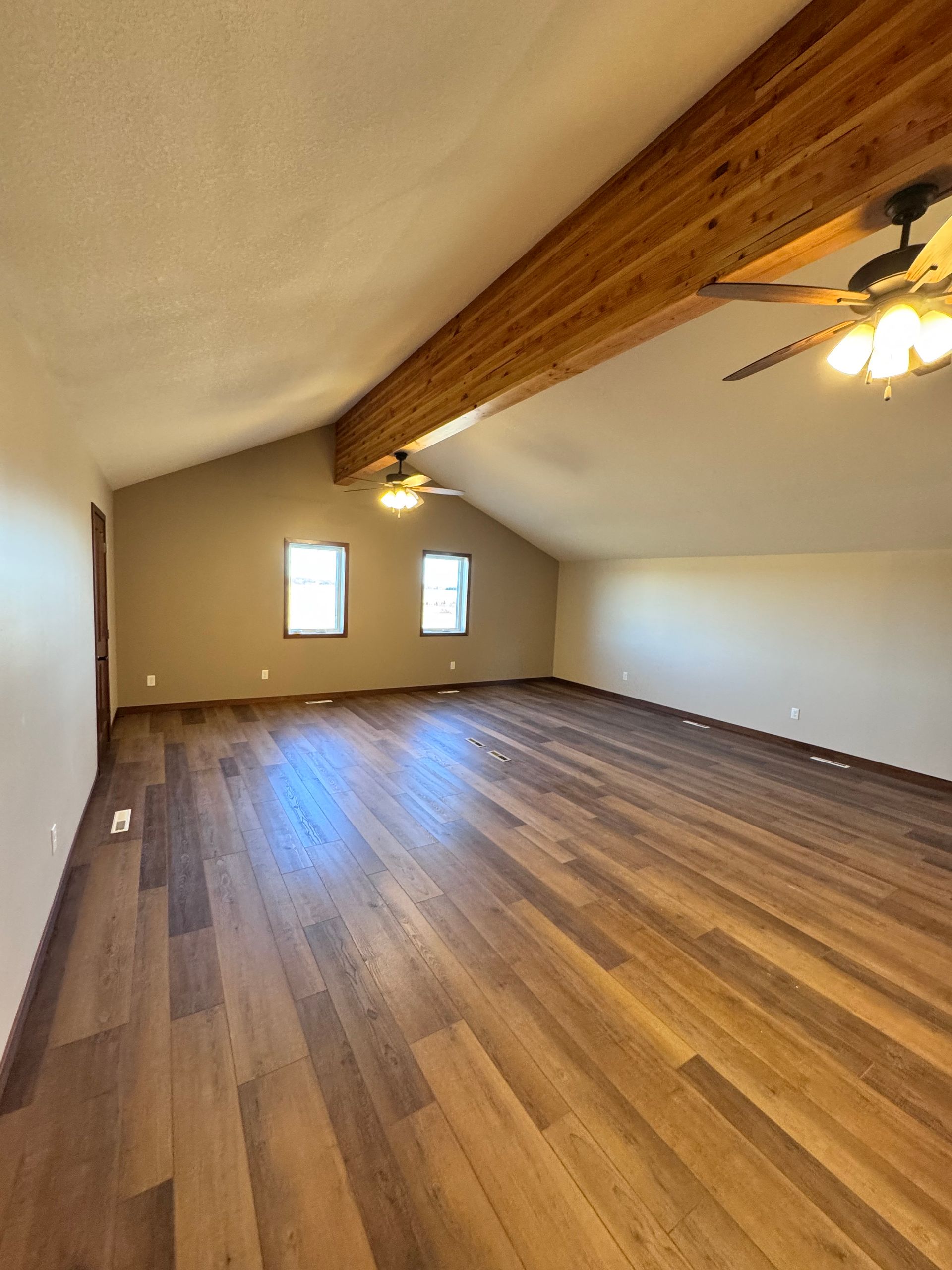Spacious room with wood floors, high ceiling with wooden beam, two windows and ceiling fans.