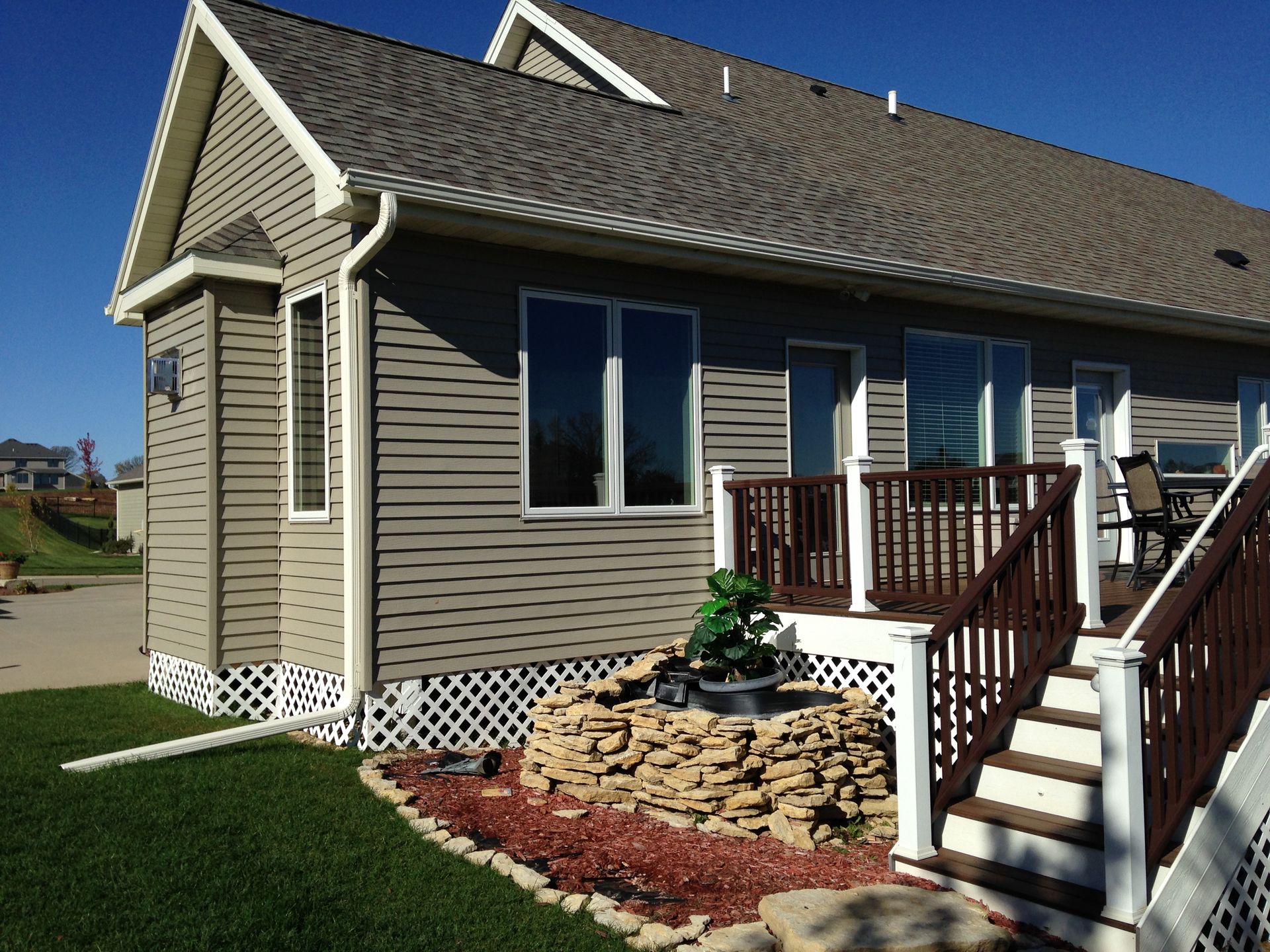 Tan house with deck, windows, and a rock-edged water feature. Green lawn, brown deck, blue sky.