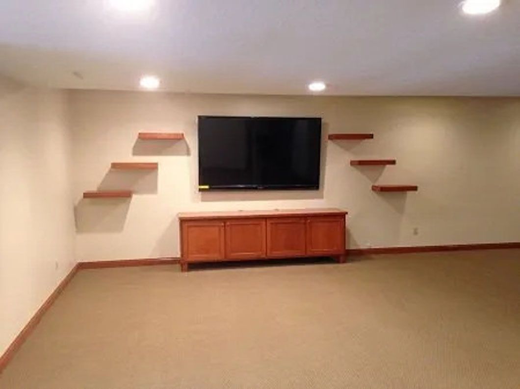 Beige room with TV, wood cabinet, and floating shelves on a wall with carpet floor.