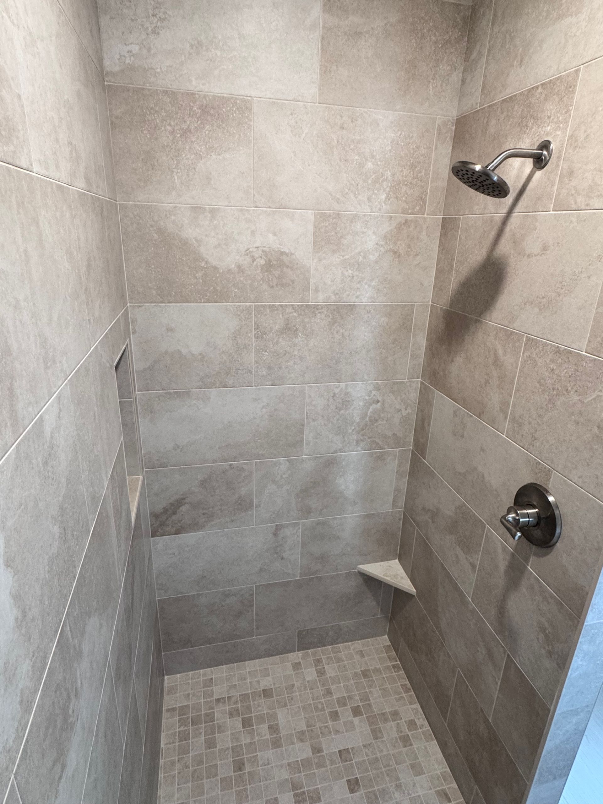 Shower with beige tiled walls, pebble floor, showerhead and control. Small corner seat.