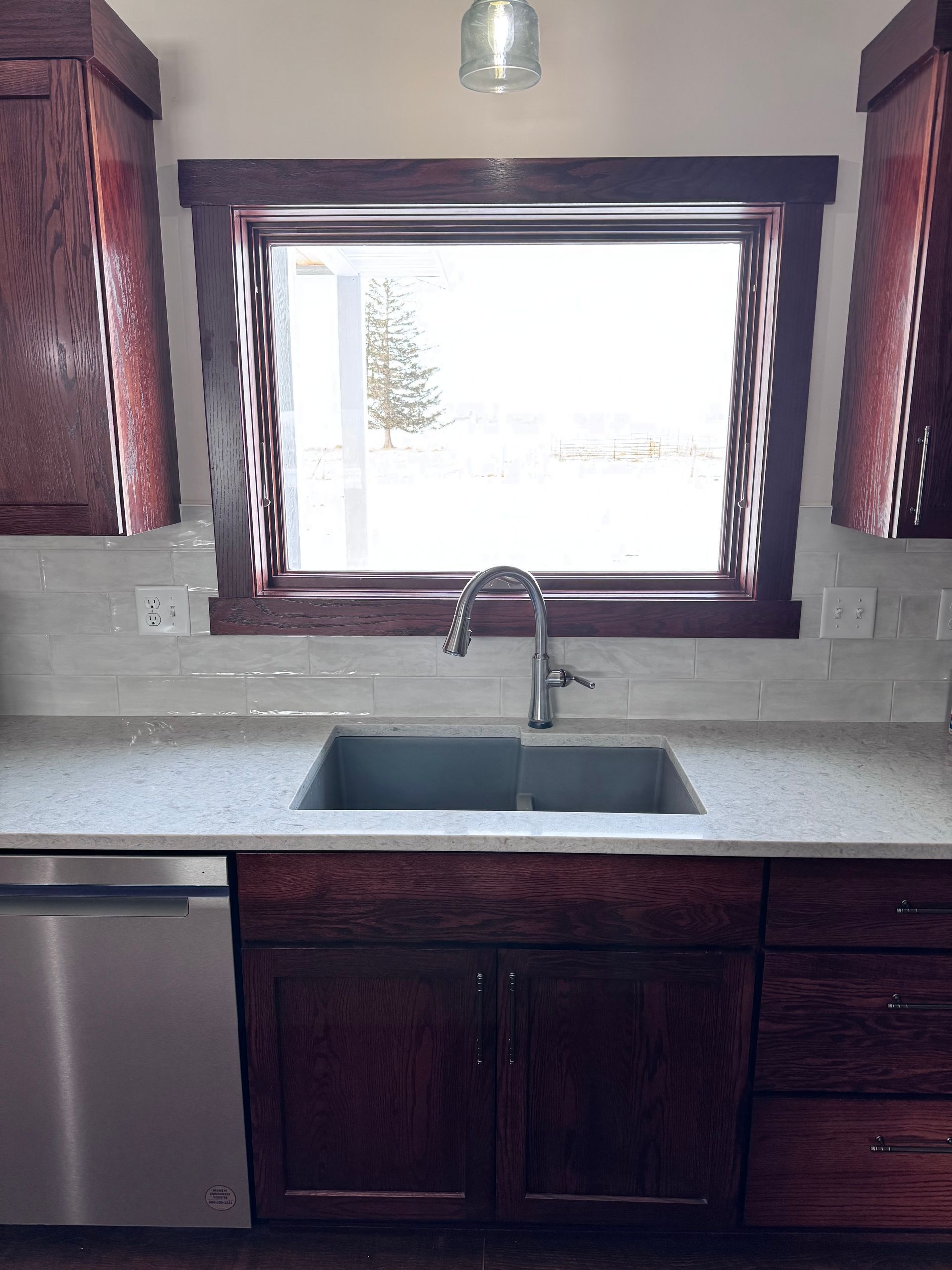 Kitchen sink with window above, dark cabinets, stainless steel appliances, and light countertops.