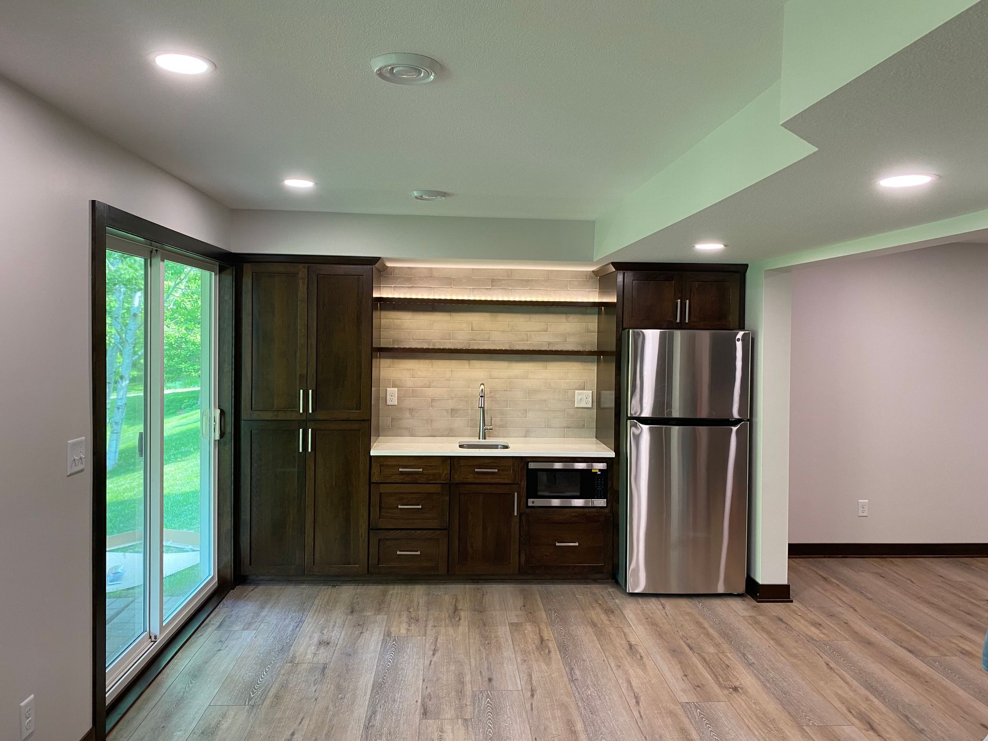 Dark wood kitchenette with stainless steel fridge, sink, microwave, and shelves. Sliding glass door to the left.