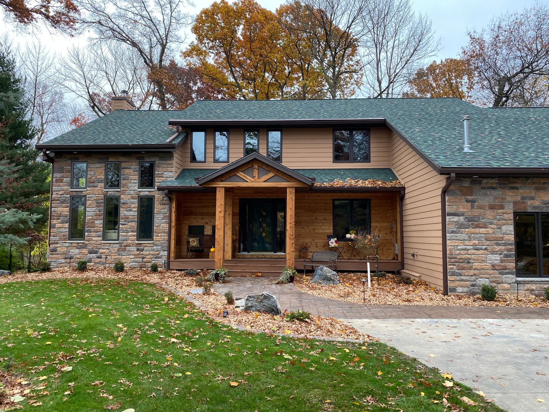 Two-story house with stone and brown siding, green roof, covered porch, and fall foliage.