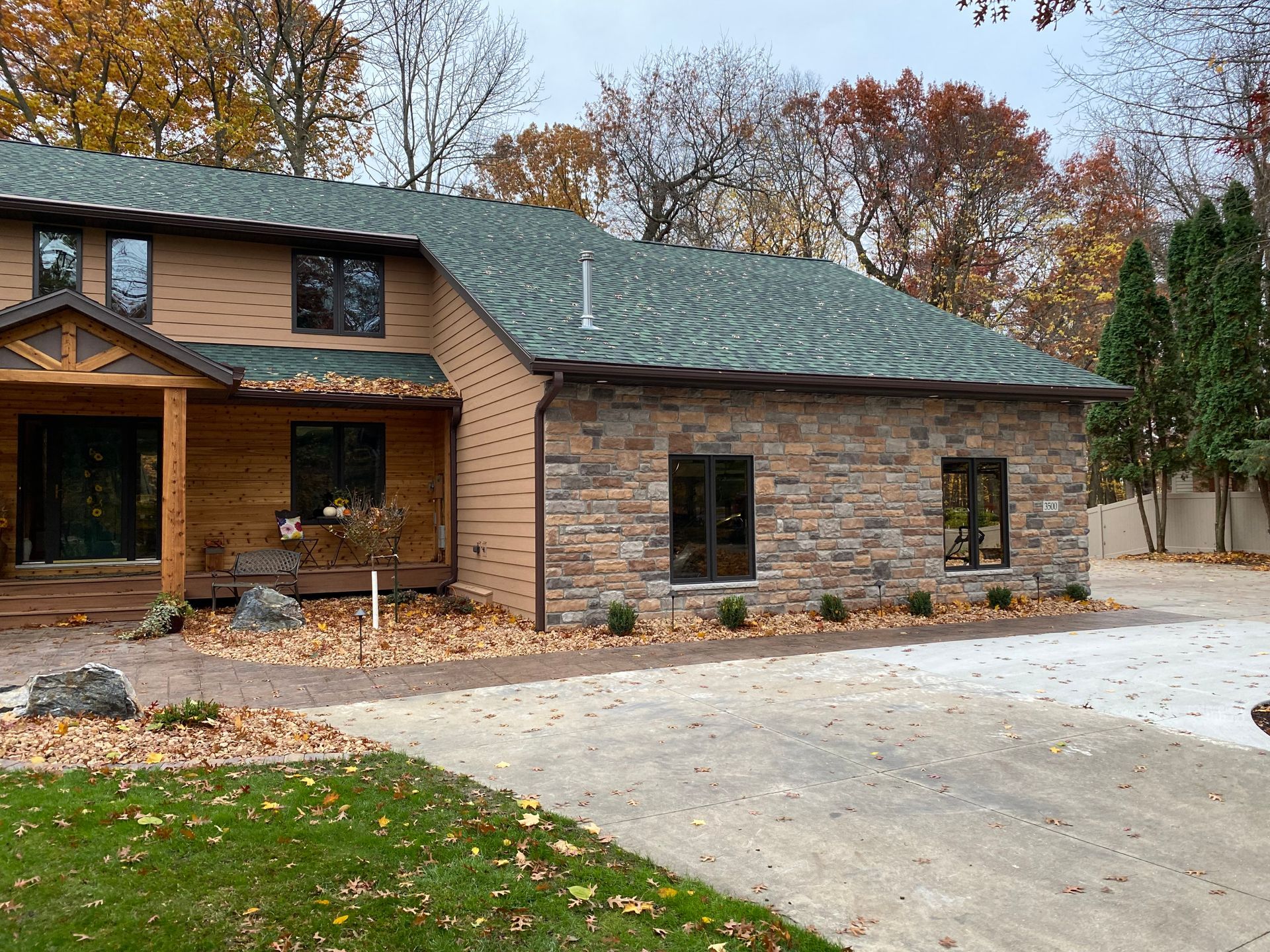 Two-story house with green roof and stone facade, surrounded by trees and a gravel driveway.
