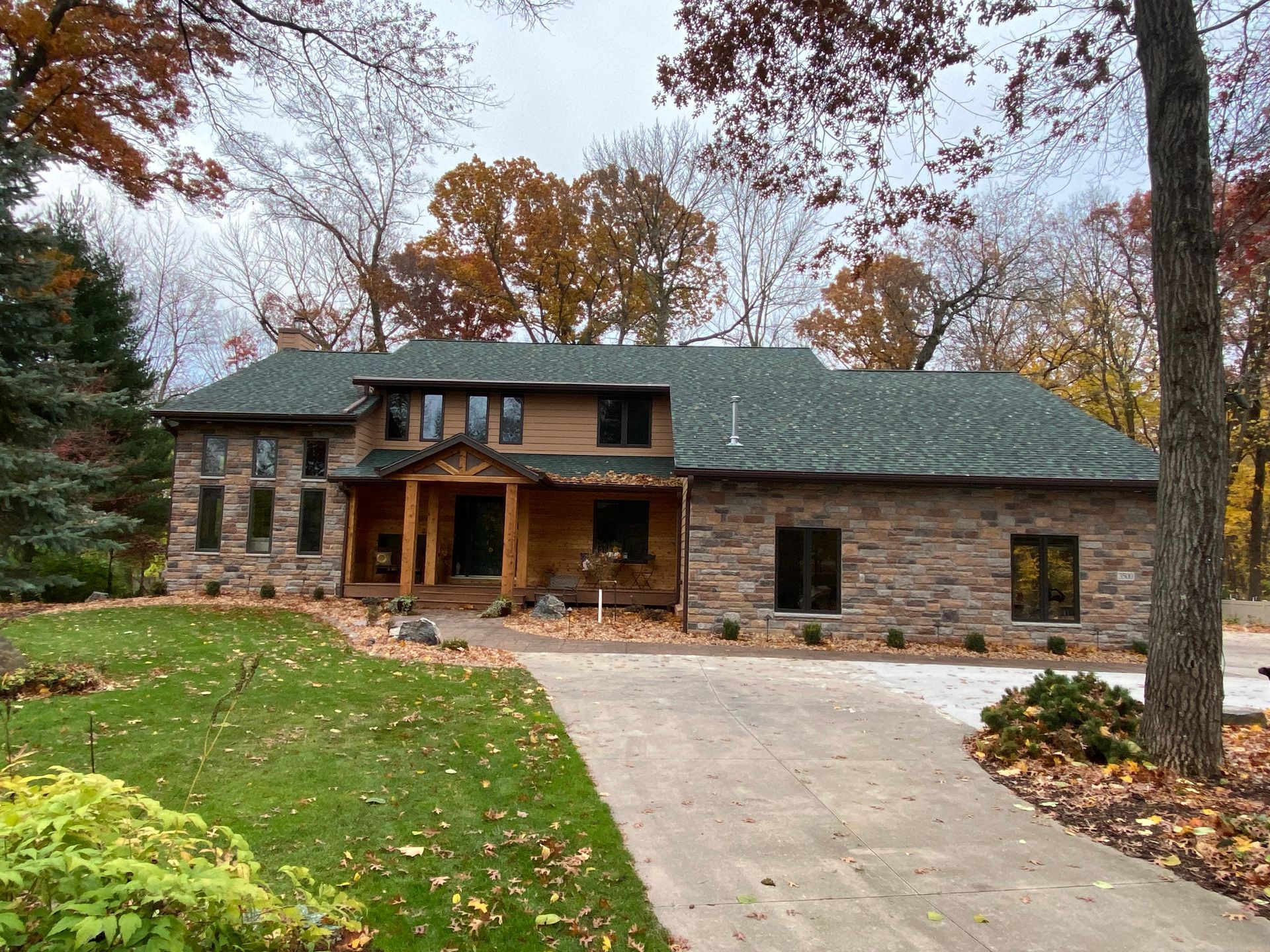 Two-story stone house with a green roof, surrounded by trees with autumn leaves and a paved driveway.