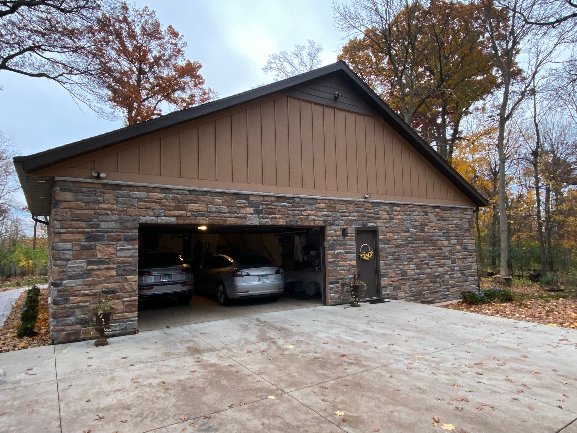 Garage with stone facade, two cars inside. Brown siding above, brown door on the right. Concrete driveway, trees in the background.