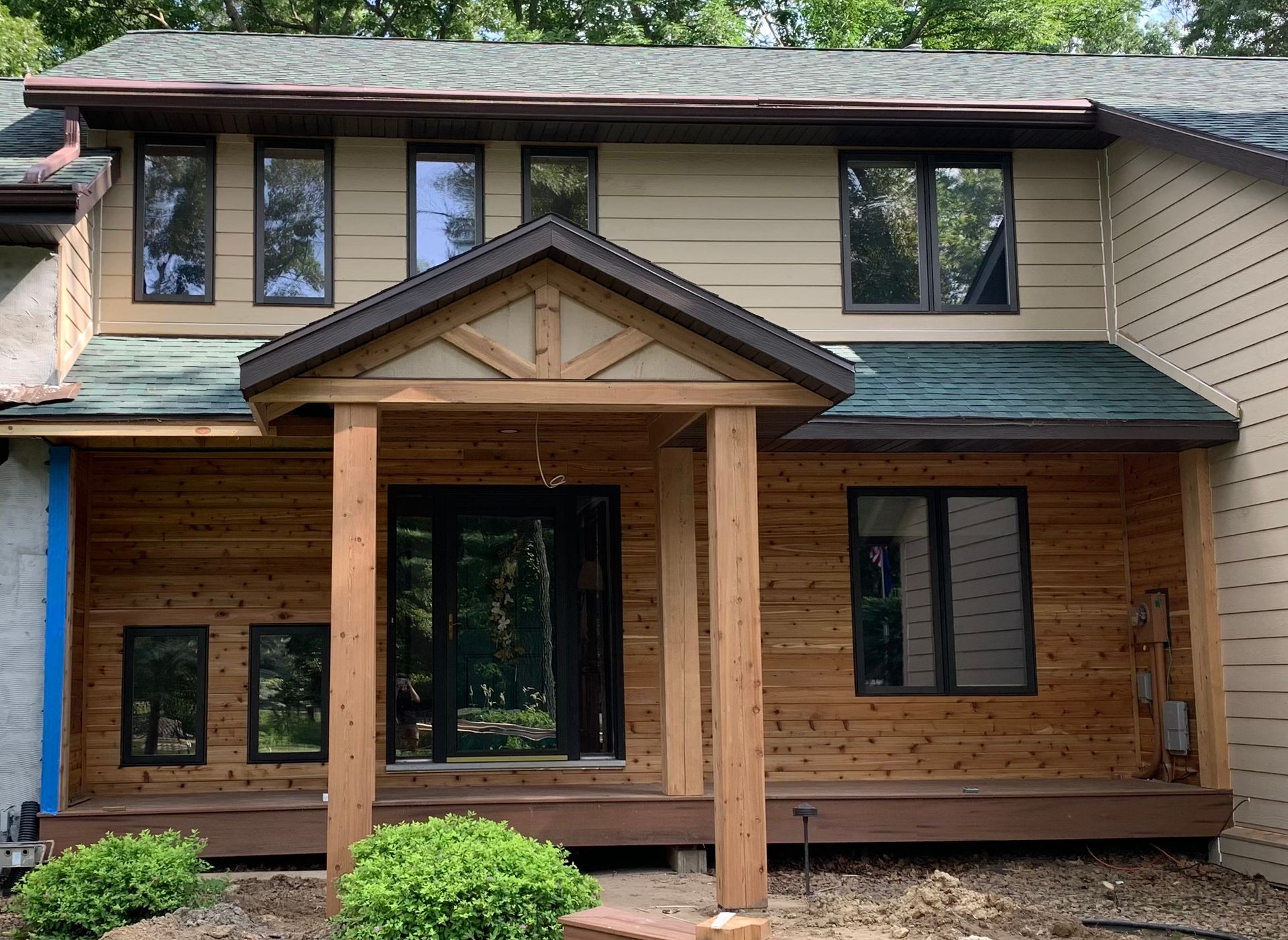 Tan and wood-paneled house with brown trim, windows, and front porch. Green roof, bushes, and visible front door.