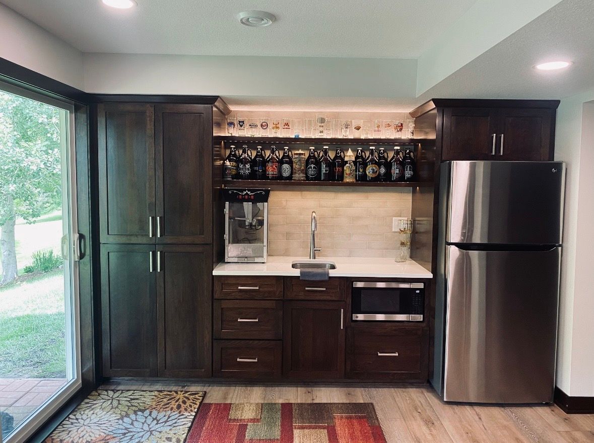 Dark-stained home bar with cabinets, shelf of liquor bottles, microwave, sink, and stainless steel refrigerator.