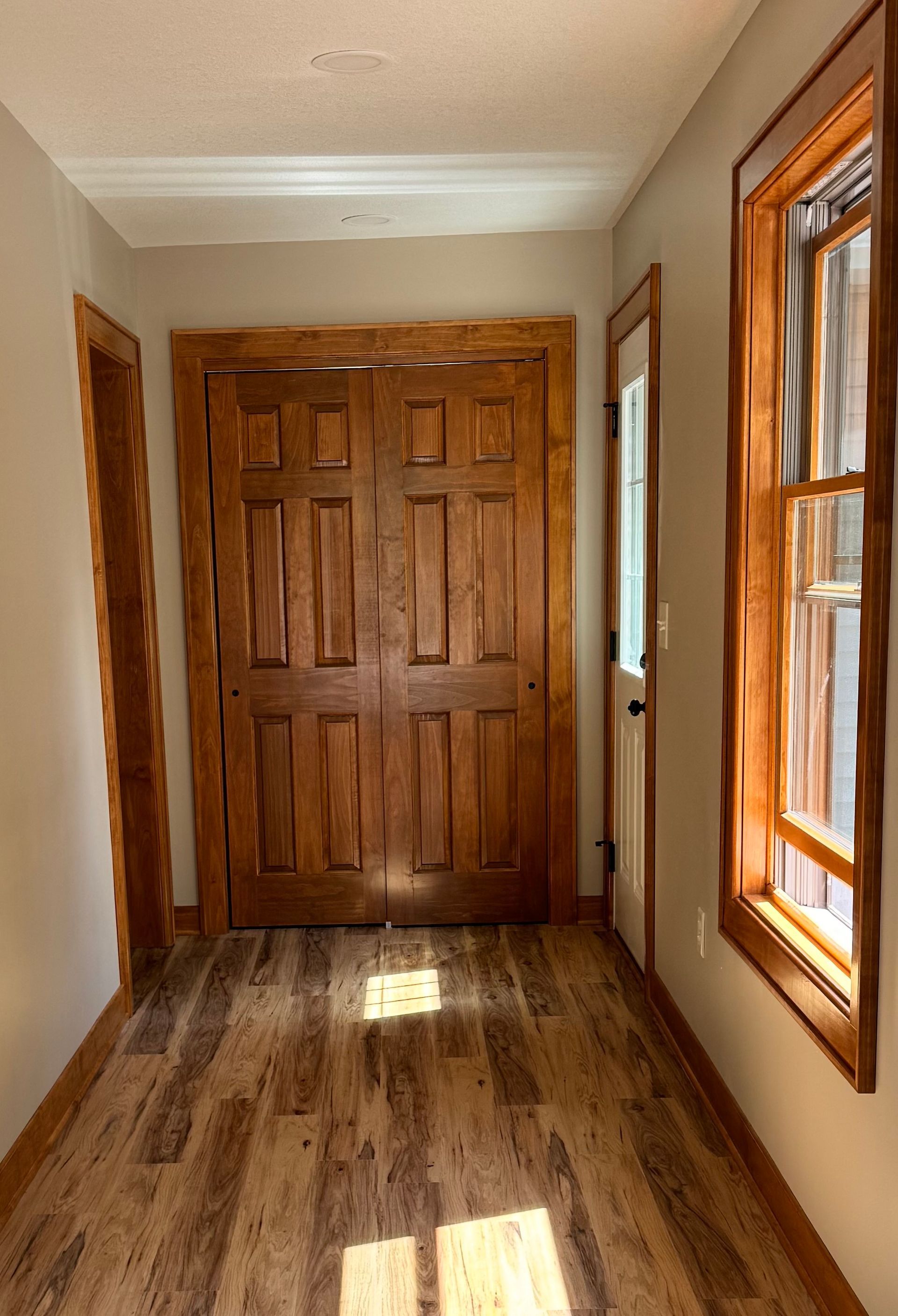 Narrow hallway with wood-paneled doors, a window, and wood-look flooring; bathed in sunlight.