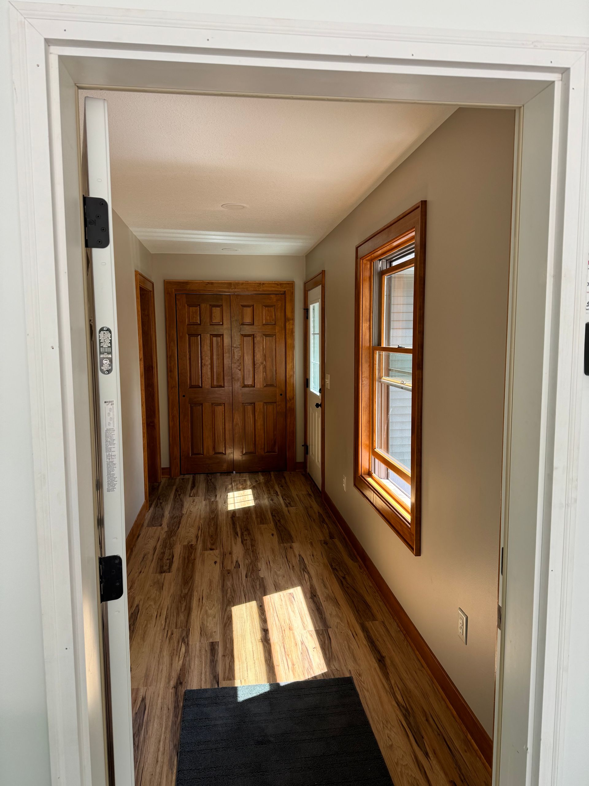 Hallway with wood floors, wood door, and a window with brown trim; lit by sunlight.