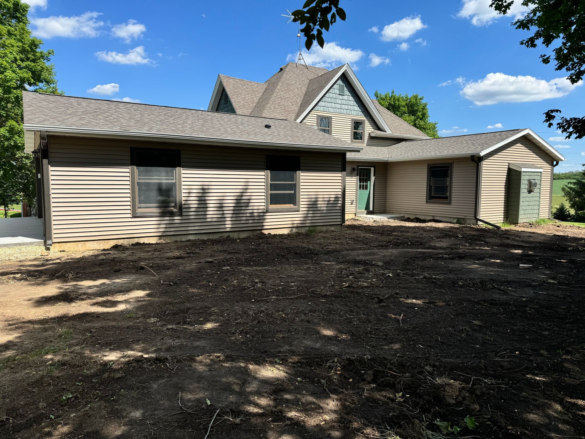 House with brown siding and roof, blue trim, and a freshly tilled yard under a blue sky with clouds.