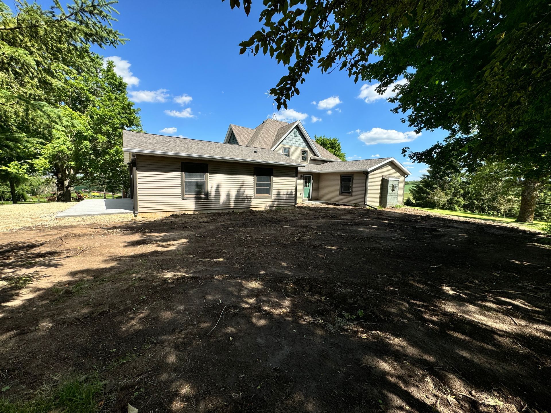 A house with a brown roof and tan siding sits on a cleared, dirt yard under a blue sky.