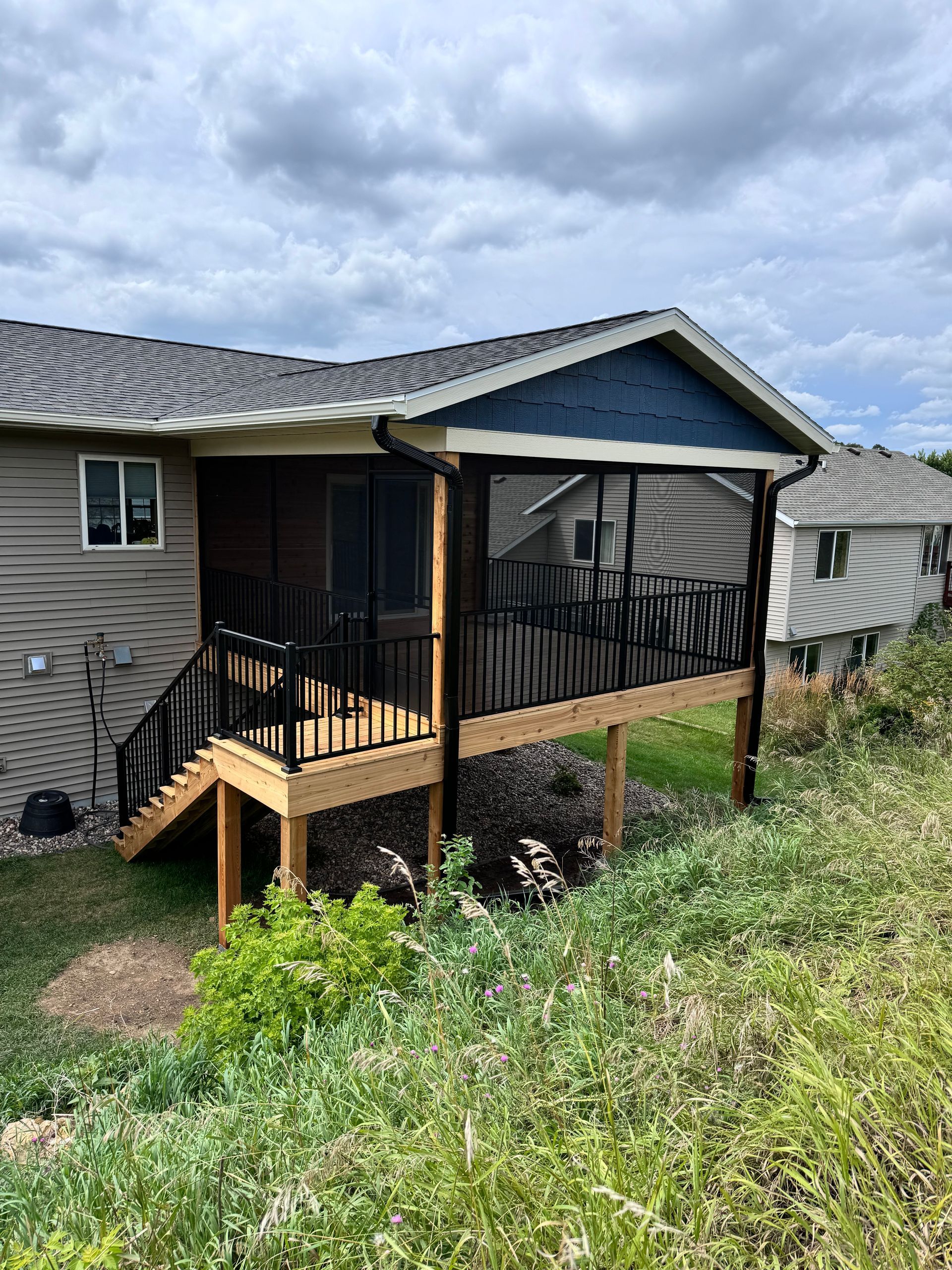 Screened porch with black railing and stairs attached to a house with tan siding, on a grassy hill.