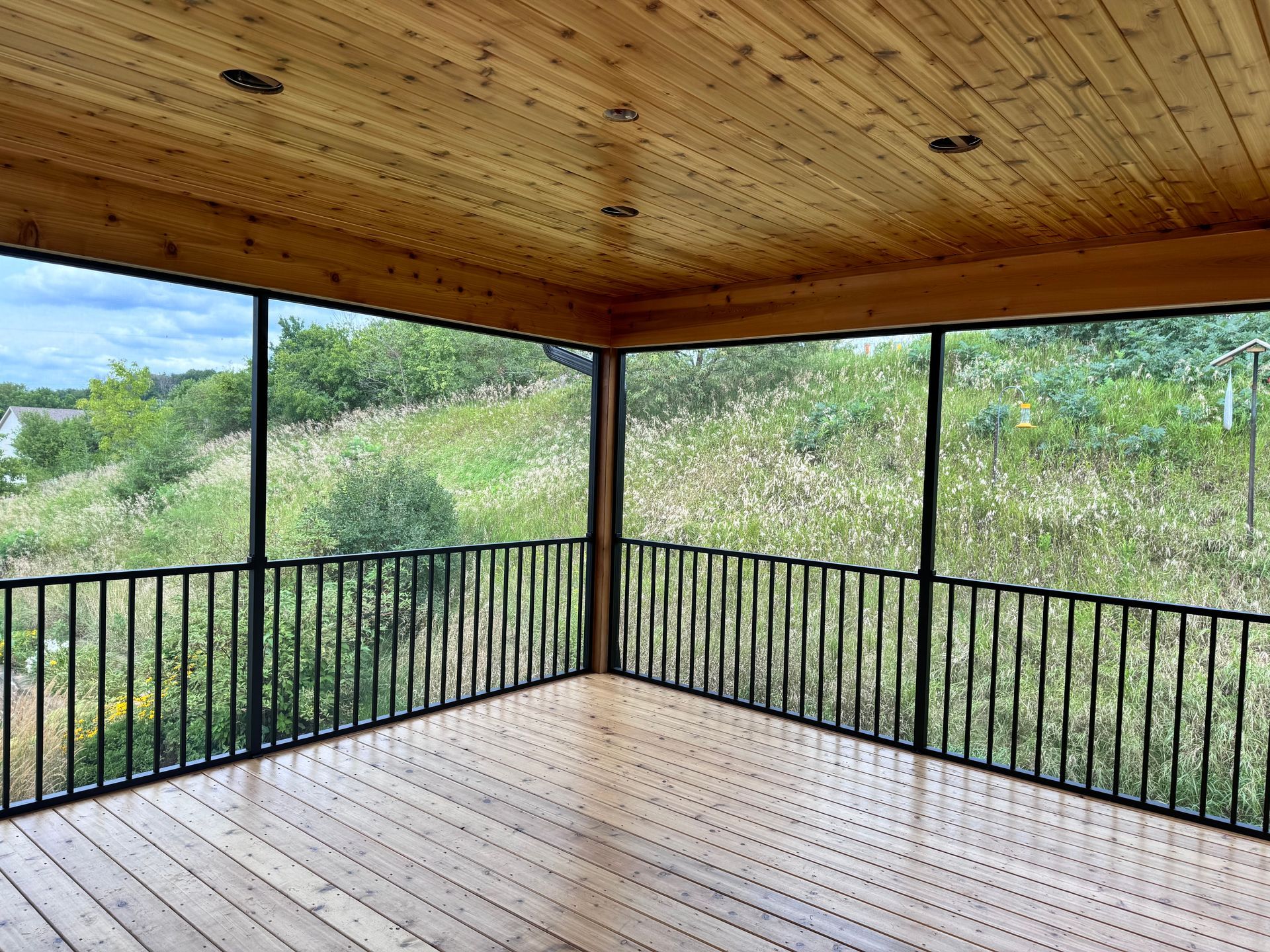 Screened-in porch with wooden floor and ceiling overlooking green hillside, framed by black railings.