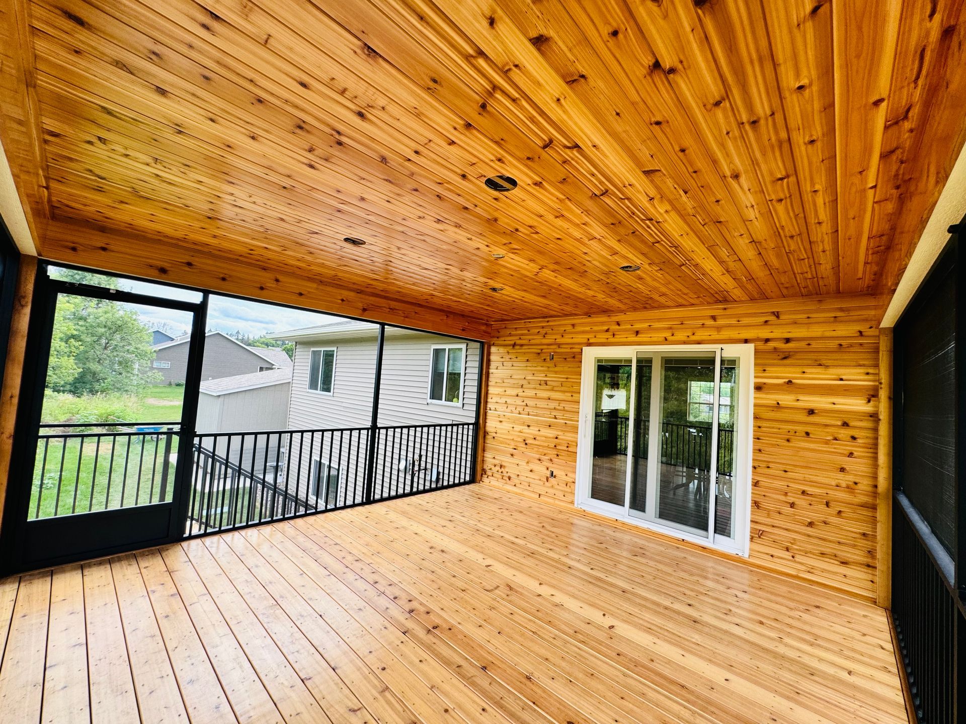 Screened-in porch with cedar wood ceiling and floor, black railings, and a glass sliding door.