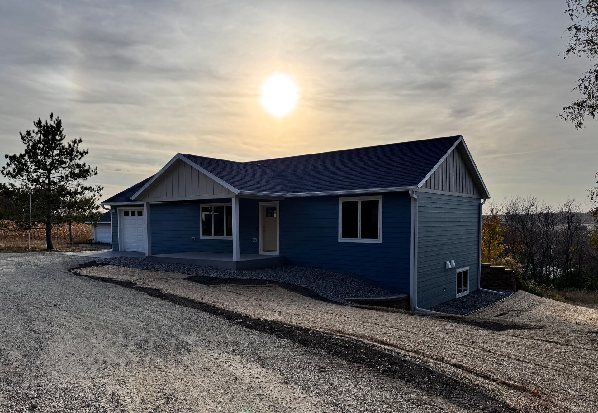 Blue house with garage, gravel driveway, and setting sun.