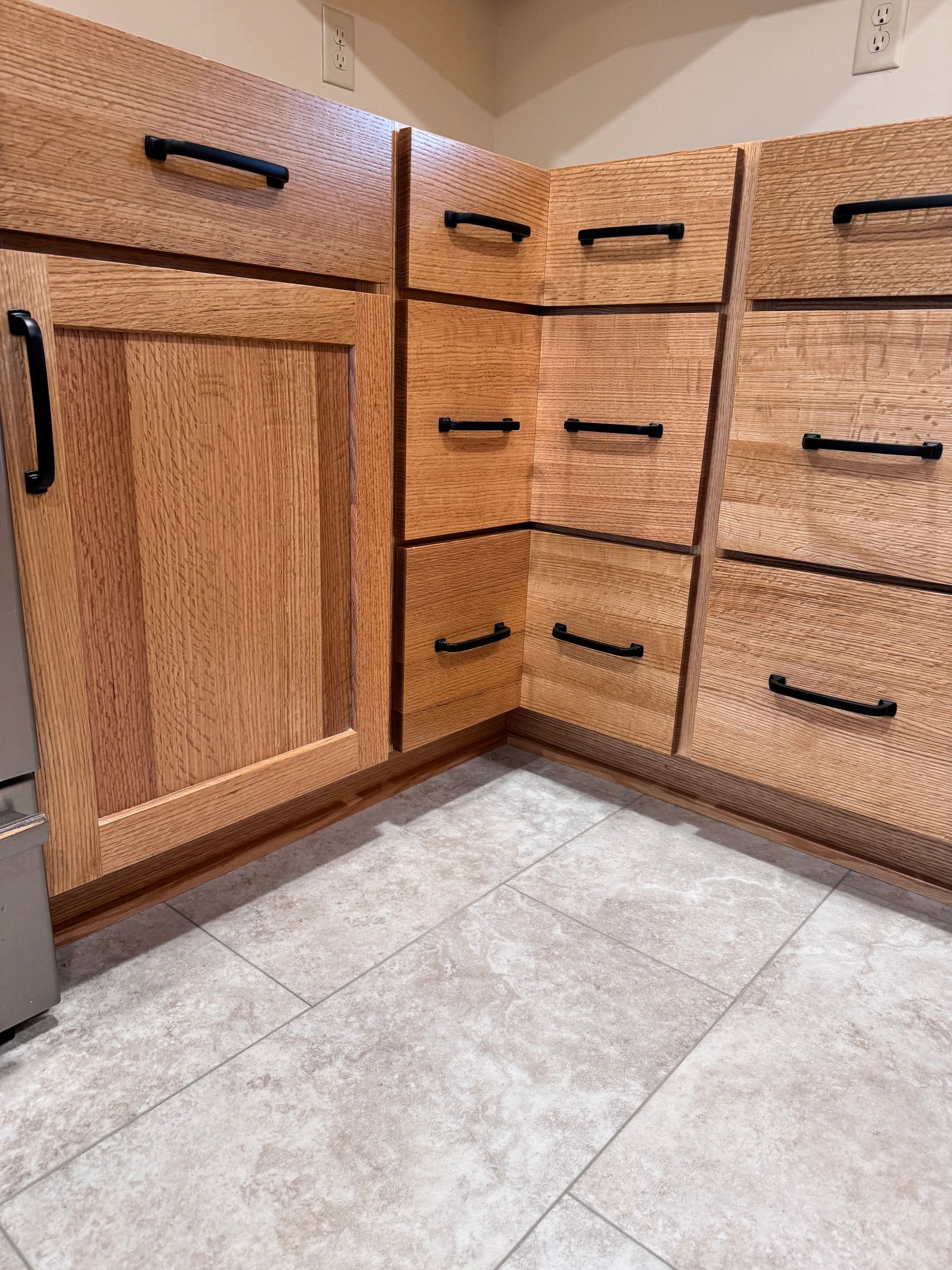 Wooden kitchen cabinets with black handles. Light wood, beige tile floor. Corner setting.
