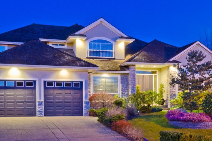 A large, well-lit house with two garage doors, a driveway, and landscaping; twilight setting.