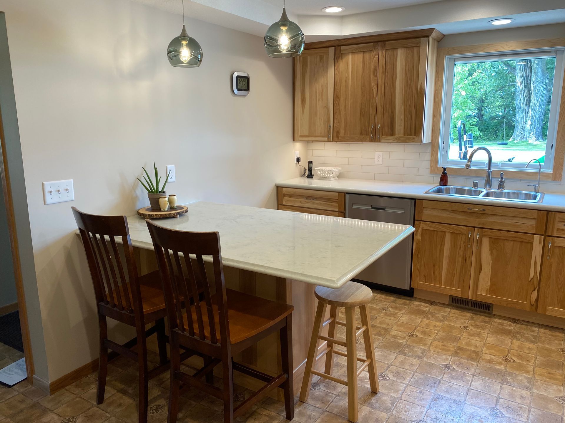 Kitchen with light wood cabinets, white countertop, two brown chairs, and a stool; a window overlooks trees.