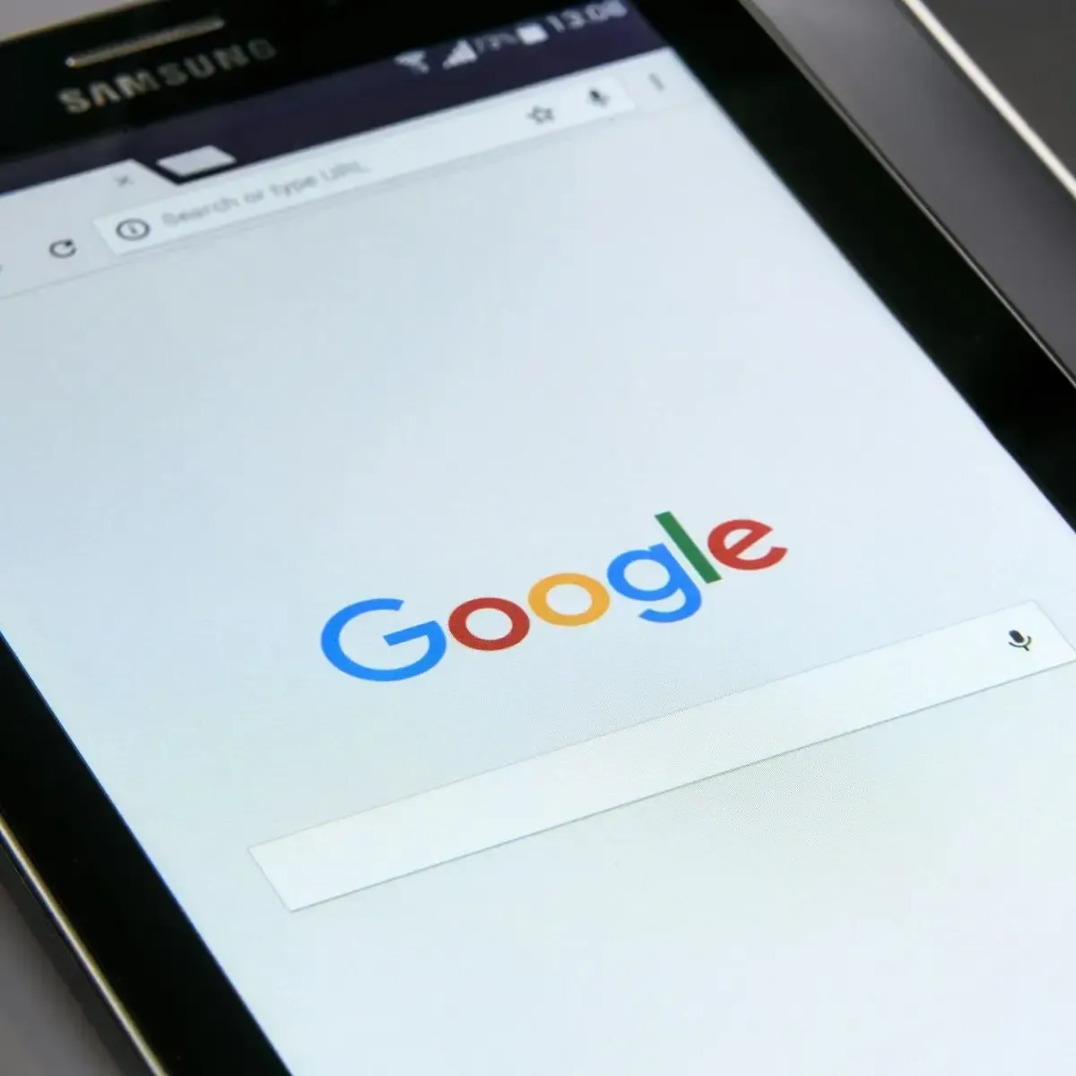 A close-up shot of a Samsung tablet displaying the Google search engine homepage, suggesting a potential tool for Google Ads management. The prominent Google logo is centered above a search bar, and a microphone icon for voice search is visible on the right side of the screen. The web browser interface shows typical elements like a URL bar at the top and basic navigation icons.